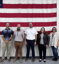 Seven individuals stand together in front of a large American flag, dressed in mixed attire, evoking patriotism and unity.