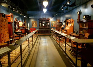 A museum exhibit features vintage furniture and artifacts against deep blue walls, creating a warm, nostalgic atmosphere.