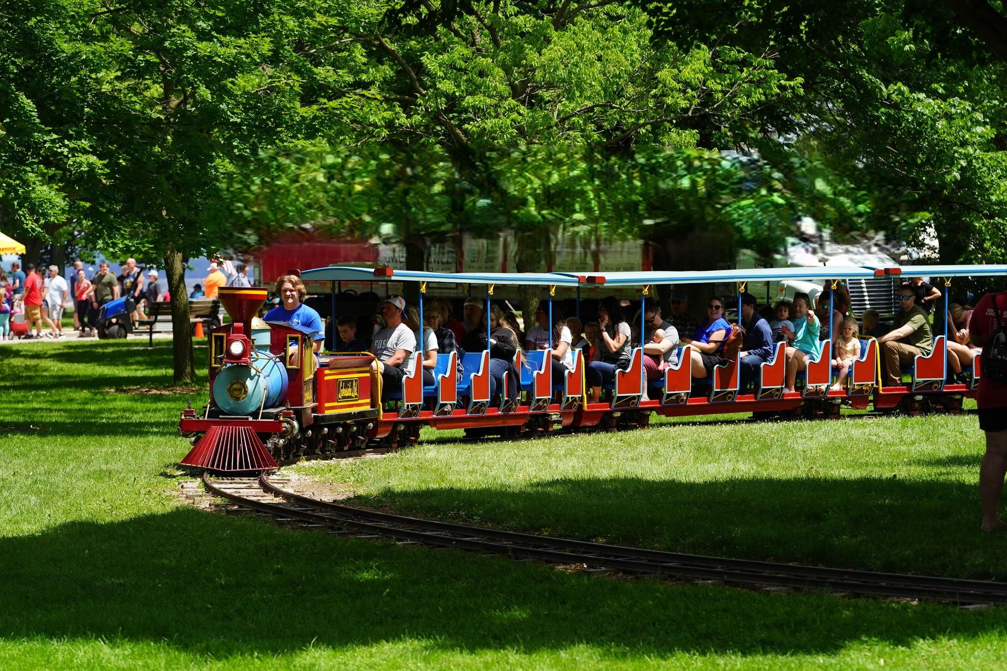 Colorful miniature train rides through a lush park, with happy people and a joyful conductor, creating a festive atmosphere.