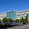 Modern multi-story hospital with glass and brick, surrounded by greenery and a busy parking lot on a clear, bright day.