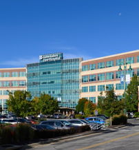 Modern multi-story hospital with glass and brick, surrounded by greenery and a busy parking lot on a clear, bright day.