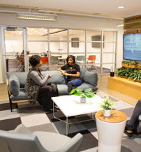 A modern lounge with comfy seating, two people chatting on a couch, and one on a phone, surrounded by greenery, fostering a friendly vibe.