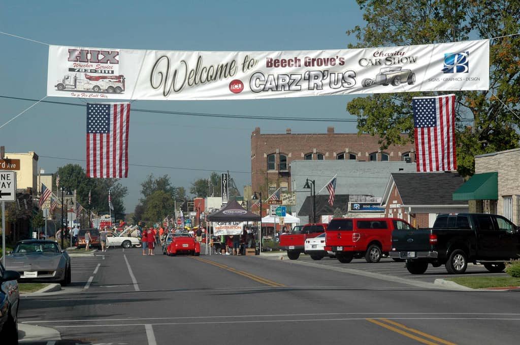 Lively street scene with a "Welcome" banner, American flags, cars, booths, and a festive, small-town atmosphere.