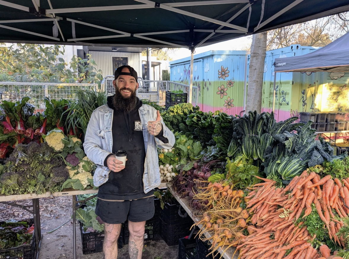 A man at a farmer's market poses with fresh vegetables, giving a thumbs-up, exuding warmth and enthusiasm for healthy living.