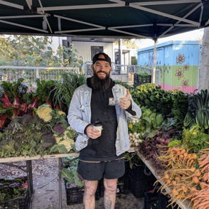 A man at a farmer's market poses with fresh vegetables, giving a thumbs-up, exuding warmth and enthusiasm for healthy living.