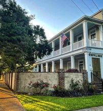 A charming two-story house with a porch, American flag, lush yard, and trees, evoking warmth and suburban peace.