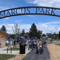 Entrance to Marcus Park, lively with families, play structures, green trees, and a bright blue sky, evoking joy and community.