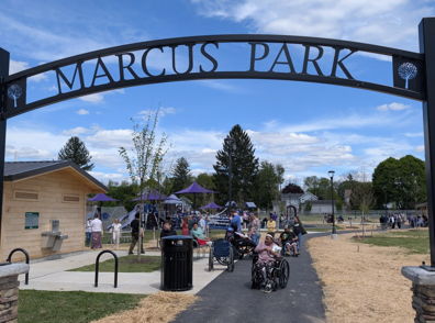 Entrance to Marcus Park, lively with families, play structures, green trees, and a bright blue sky, evoking joy and community.