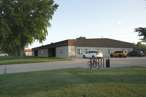 A calm, inviting scene of a modest building, vehicles, secured bicycles, green grass, and trees under a clear sky.