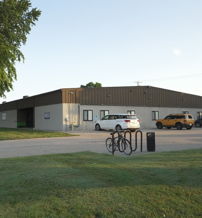 A calm, inviting scene of a modest building, vehicles, secured bicycles, green grass, and trees under a clear sky.