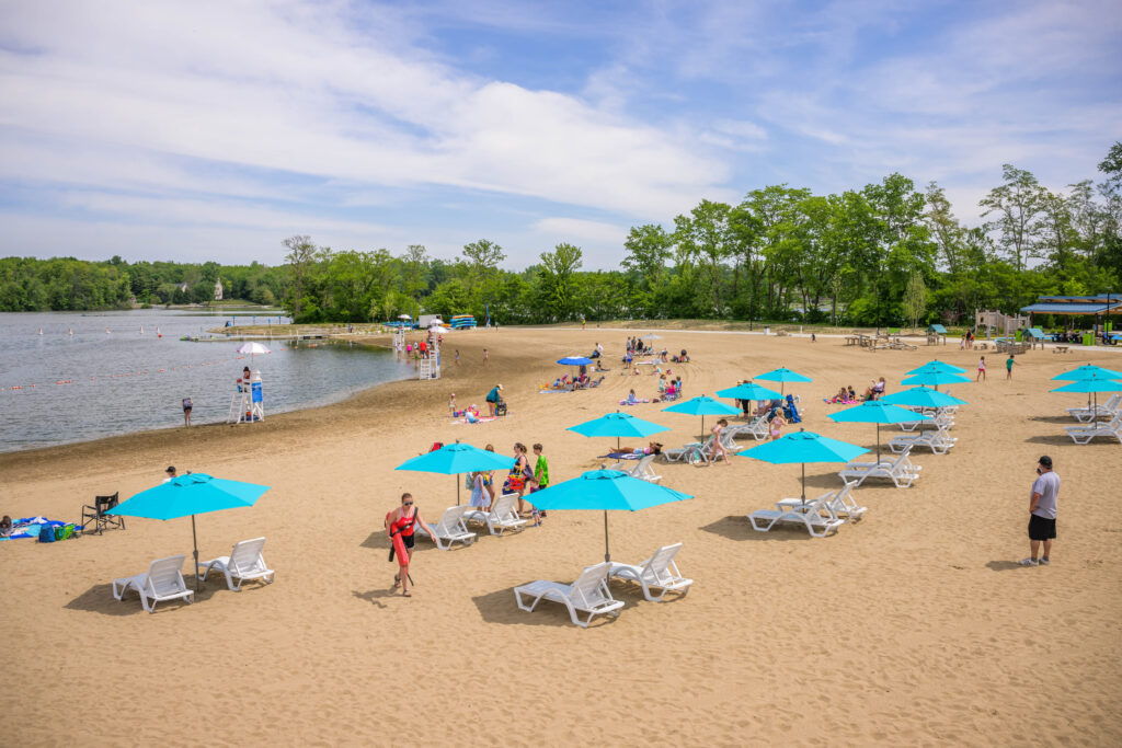 Bright beach scene with turquoise umbrellas, lounge chairs, and people enjoying sunbathing, swimming, and socializing.