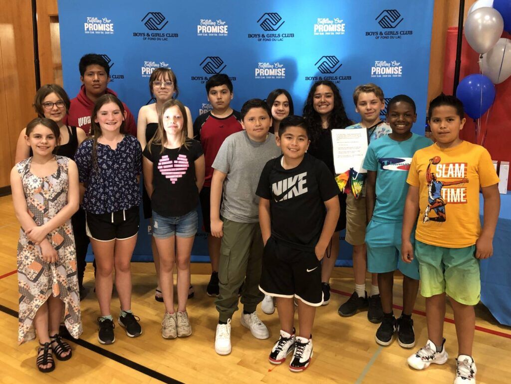 A joyful group of diverse kids and teens smiles in a gym, celebrating together under a blue backdrop with balloons.