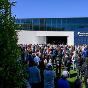 A lively crowd gathers outside a modern arts venue on a sunny day, filled with excitement, joy, and community anticipation.