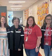 Five smiling women in casual attire stand in a bright hallway, showcasing a vibrant, supportive atmosphere.