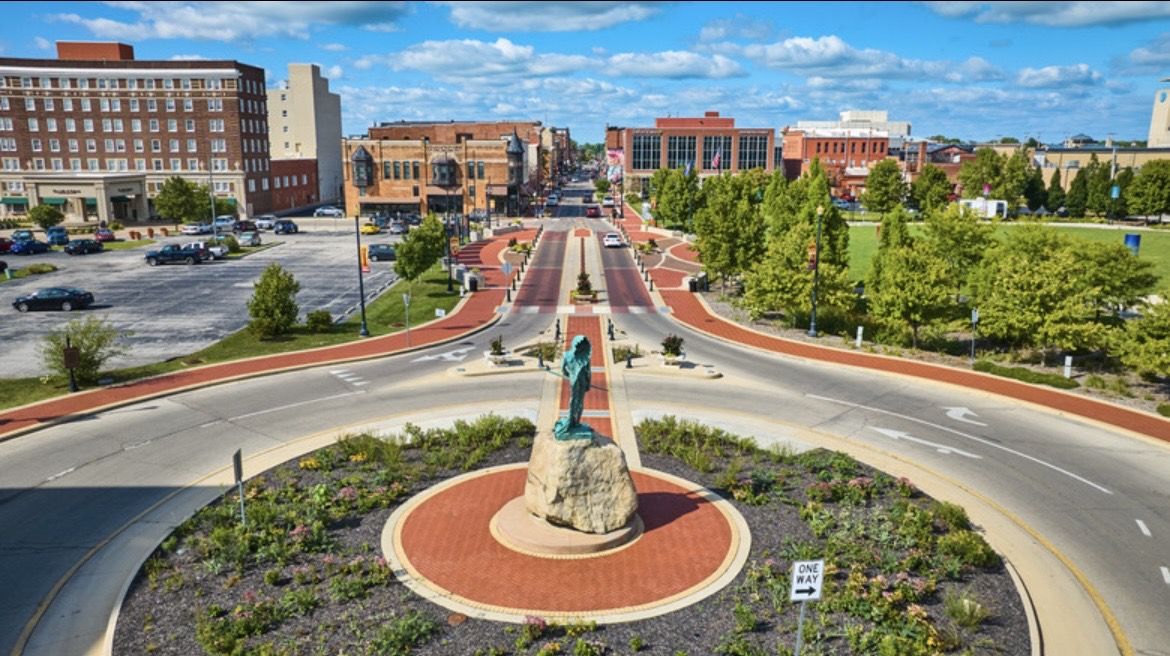 A vibrant urban roundabout with a large statue, surrounded by flowers, buildings, trees, and blue skies, exudes liveliness and serenity.
