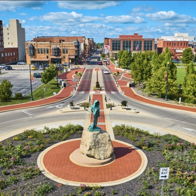 A vibrant urban roundabout with a large statue, surrounded by flowers, buildings, trees, and blue skies, exudes liveliness and serenity.