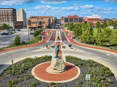 A vibrant urban roundabout with a large statue, surrounded by flowers, buildings, trees, and blue skies, exudes liveliness and serenity.