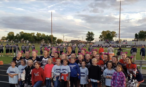 Cheerleaders and kids pose joyfully on a football field at a community event, promoting cheer and support, with a cloudy sky.