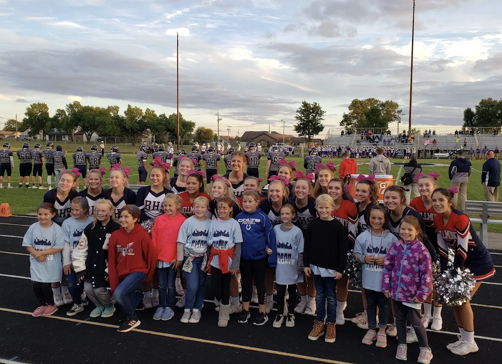 Cheerleaders and kids pose joyfully on a football field at a community event, promoting cheer and support, with a cloudy sky.