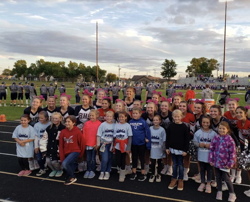 Cheerleaders and kids pose joyfully on a football field at a community event, promoting cheer and support, with a cloudy sky.