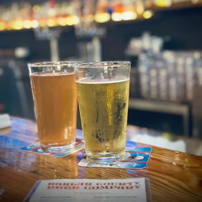 Two glasses of beer on a wooden bar, surrounded by softly lit shelves, creating a warm, inviting pub atmosphere.