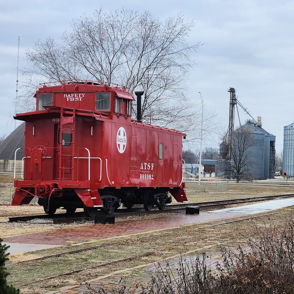 A vibrant red caboose on railway tracks contrasts with gray silos under a cloudy sky, evoking nostalgia and tranquility.