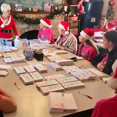 A group of people in red hats gathers around a table with envelopes, engaged in festive holiday preparations, sharing joy together.