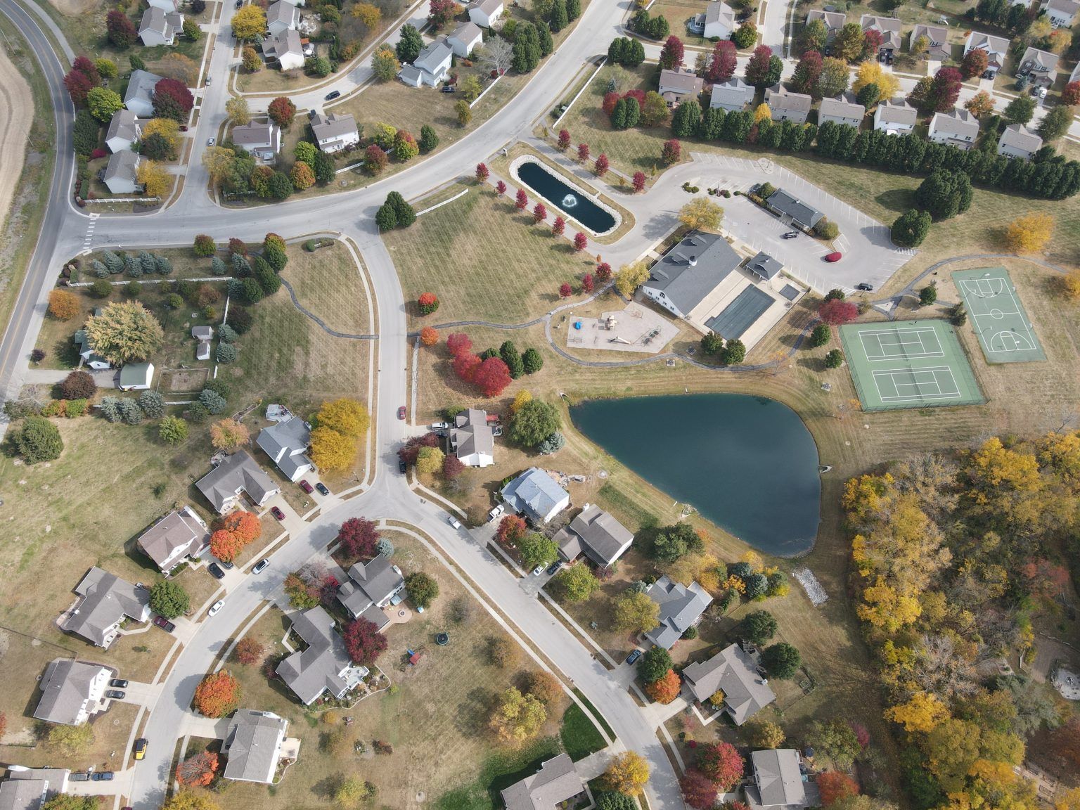 Aerial view of a suburban neighborhood with houses, winding roads, autumn foliage, a serene pond, and tennis courts.