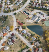 Aerial view of a suburban neighborhood with houses, winding roads, autumn foliage, a serene pond, and tennis courts.