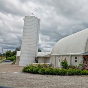 A barn-like building with a curved roof and silos amidst greenery, exuding rustic charm under an overcast sky.