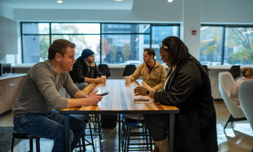 A modern cafe scene with five people at a table; some are focused on devices, others engage in conversation.