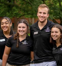 Five smiling individuals in matching black polo shirts stand together outdoors, exuding camaraderie and joy in a vibrant setting.