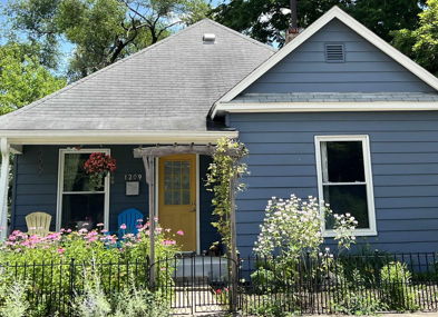 Charming blue house with sloped roof, colorful porch chairs, and vibrant flowers, creating a warm and inviting atmosphere.