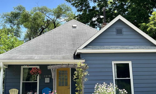 Charming blue house with sloped roof, colorful porch chairs, and vibrant flowers, creating a warm and inviting atmosphere.