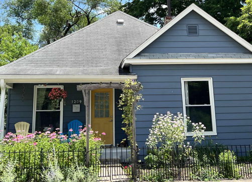 Charming blue house with sloped roof, colorful porch chairs, and vibrant flowers, creating a warm and inviting atmosphere.