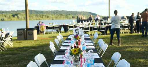 A long outdoor dining setup with white chairs, a black tablecloth, and cheerful decorations, set by a serene water backdrop.