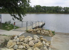 Serene lakeside view with a wooden dock, calm waters, large rocks, and greenery under an overcast sky, evoking tranquility.