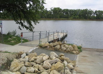 Serene lakeside view with a wooden dock, calm waters, large rocks, and greenery under an overcast sky, evoking tranquility.