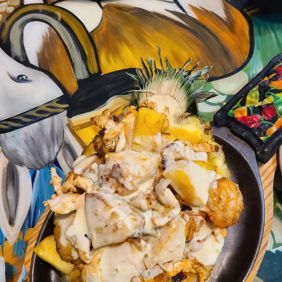 A vibrant plate features grilled chicken, diced potatoes in creamy sauce, garnished with pineapple, against a colorful mural backdrop.