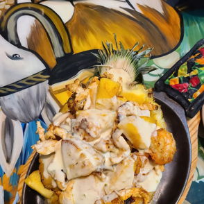 A vibrant plate features grilled chicken, diced potatoes in creamy sauce, garnished with pineapple, against a colorful mural backdrop.