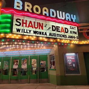 Vintage theater marquee in neon lights displays "SHAUN OF THE DEAD" and auditions for "WILLY WONKA," evoking nostalgia and excitement.