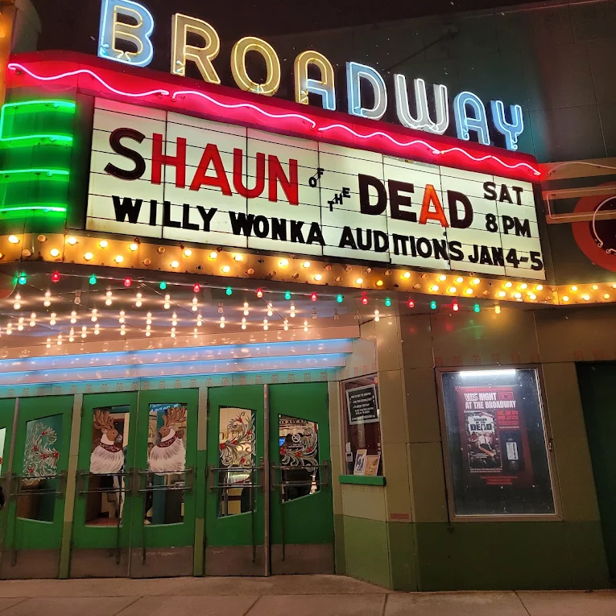 Vintage theater marquee in neon lights displays "SHAUN OF THE DEAD" and auditions for "WILLY WONKA," evoking nostalgia and excitement.