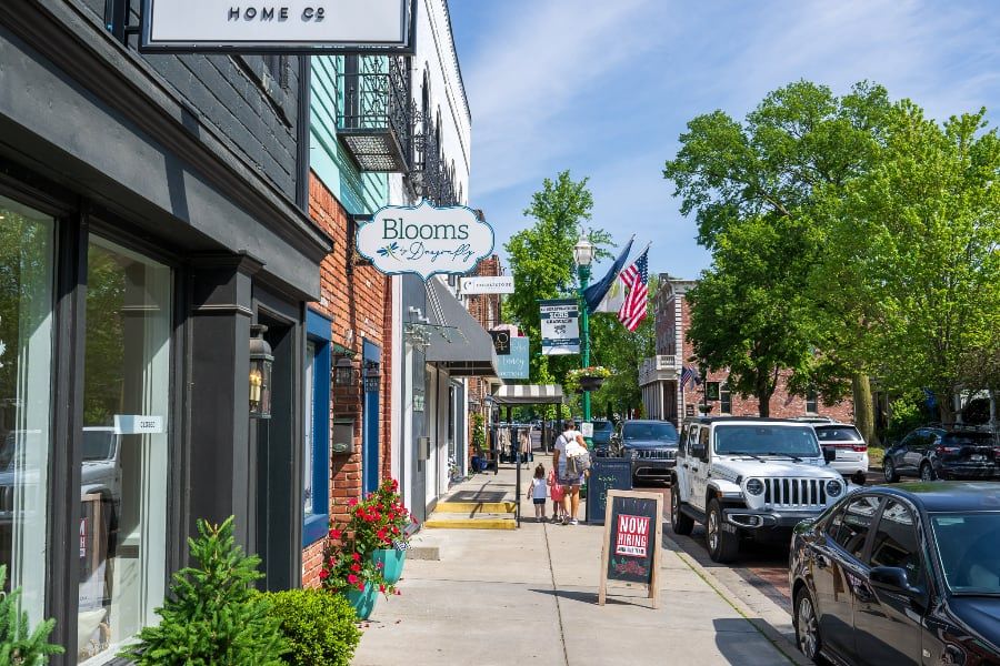 Vibrant streetscape with shops, potted plants, people strolling, flags, and sunlight, evoking warmth and community.