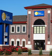 Modern bank building with a brick exterior, blue/yellow sign, surrounded by vibrant flower beds, evoking professionalism and trust.