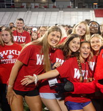 Young students in red shirts gather joyfully at a sports field, celebrating with a smiling mascot, exuding excitement and unity.