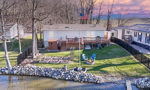 A tranquil lakeside scene features a cozy house, dock, and lounge chairs, surrounded by trees and a flagpole with an American flag.
