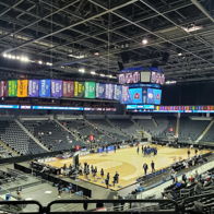 Indoor sports arena for a basketball game, with warm-up players, empty stands, colorful banners, and a large scoreboard.