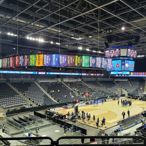 Indoor sports arena for a basketball game, with warm-up players, empty stands, colorful banners, and a large scoreboard.