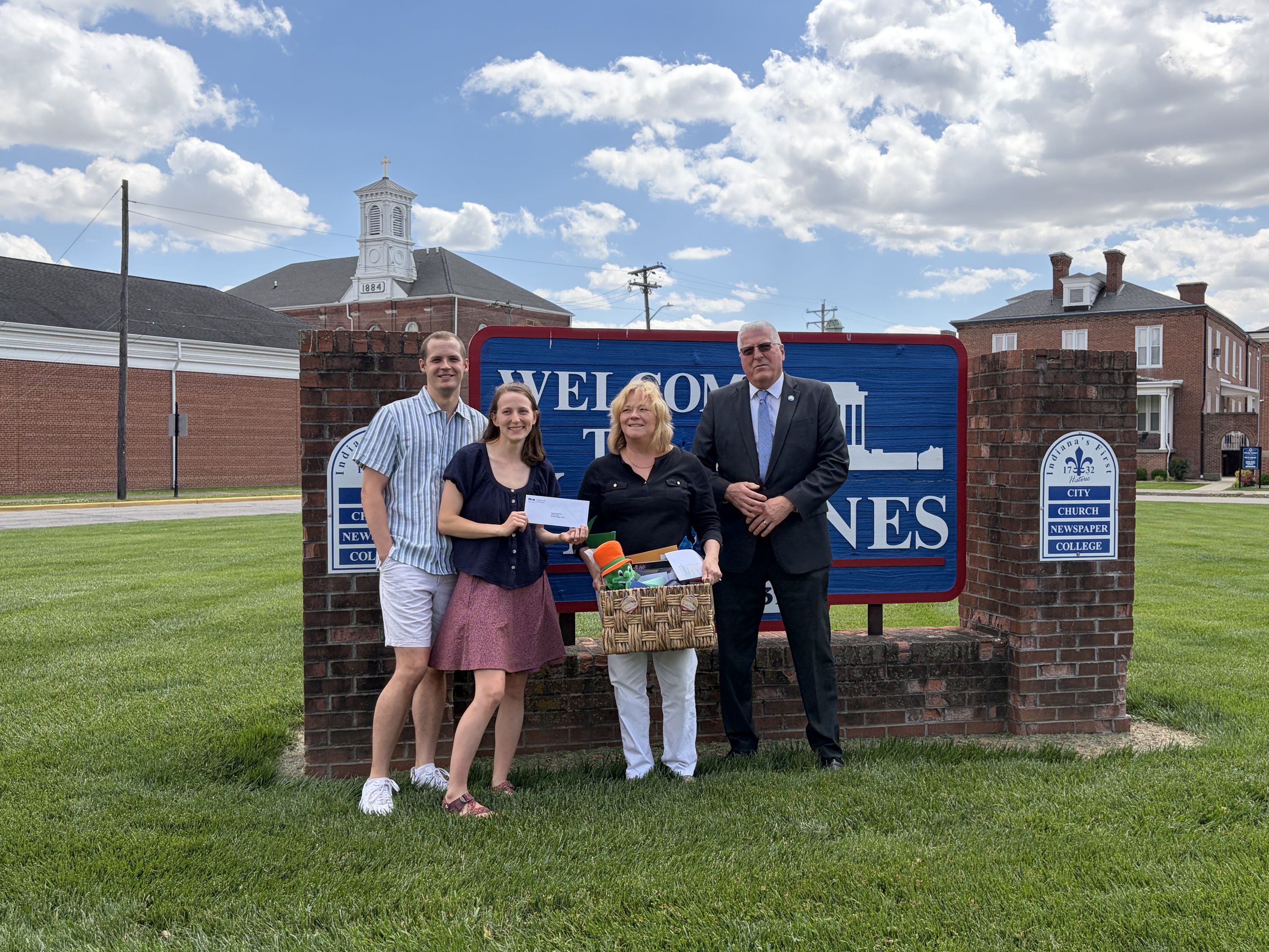 Four smiling people pose by a "Welcome to Jones" sign, with a grassy area and buildings under a bright blue sky.