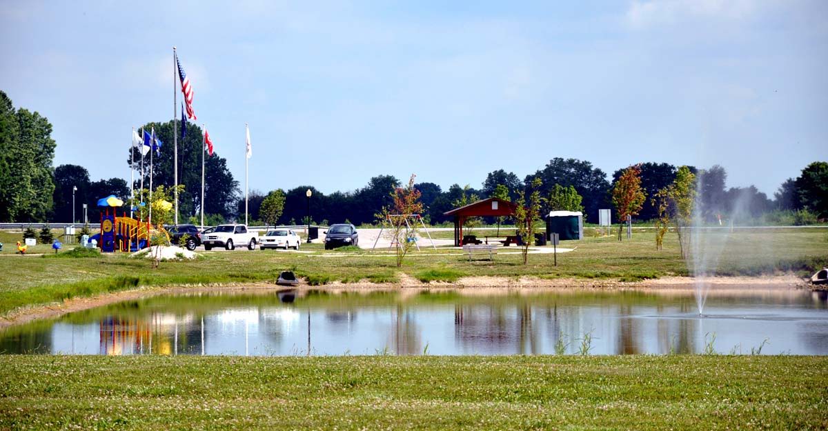 Serene park with a reflective pond, greenery, trees, playground, and shelter, evoking tranquility and community leisure.